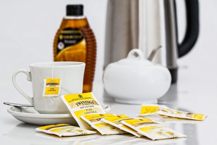 An image of a teacup with several bags of tea laying next to it and a sugar bowl, bottle of honey and a metal picture in the background